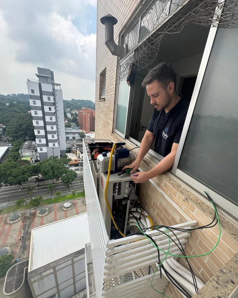 Técnico da SeuAr Climatização com a bomba de vácuo ligada na condensadora, na sacada técnica de um apartamento em São Paulo